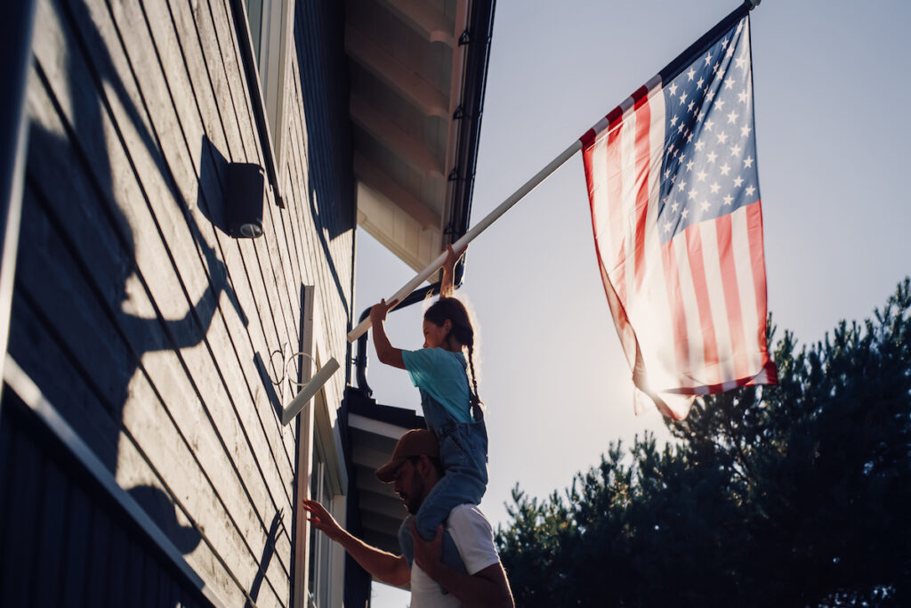 Proud Patriotic Dad Holding His Young Daughter of His Shoulders, Helping Her to Put the United States of America Flag on the Wall of Their House to Celebrate a National Holiday Sunny Day in USA