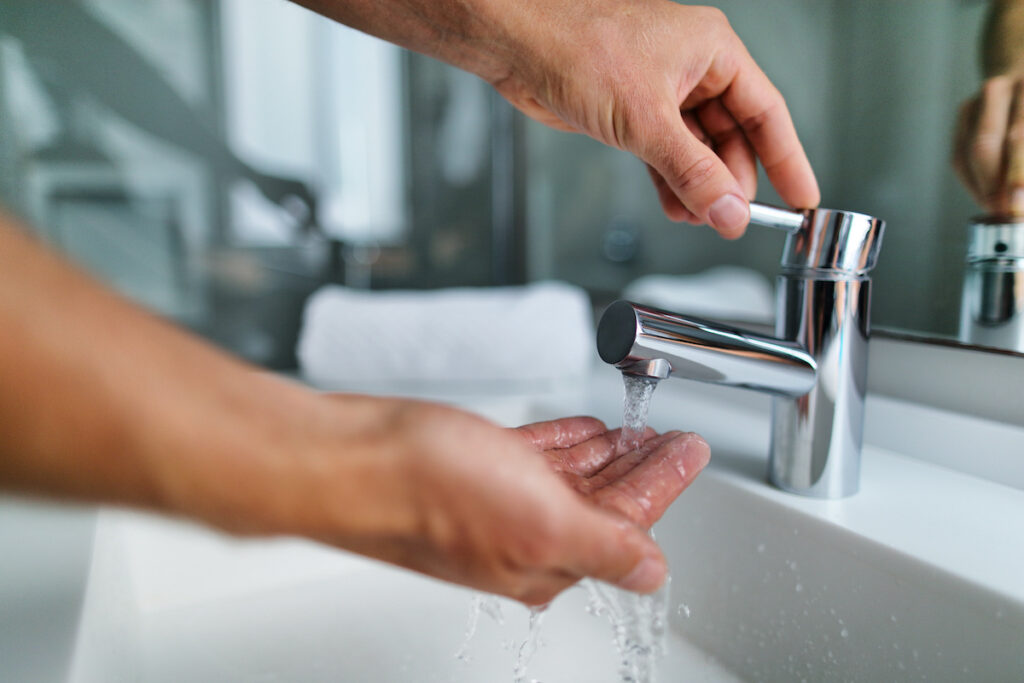 Man washing hands in bathroom sink at home checking temperature touching running water with hand Closeup on fingers under hot water out of a faucet of a sink